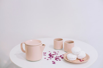 White table with a pink teapot and mugs and some tasty marshmallows on a plate in a white room
