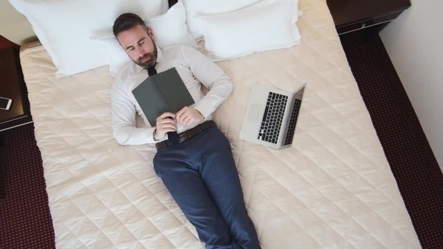 Crane shot of businessman in formalwear lying on hotel bed reading book and then falling asleep