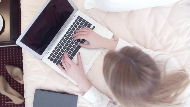 Crane shot of young businesswoman sitting on hotel bed and working on laptop