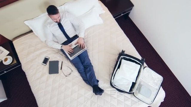 Crane shot of businesswoman in formalwear sitting on bed typing on laptop, then lying down and closing his eyes
