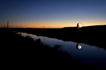 Man is dancing with fire on the ropes near water with reflections on the sunset