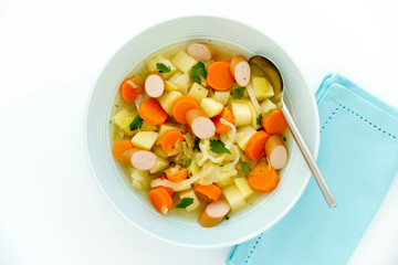 Vegetable Stew with sliced Frankfurter sausage in a blue soup bowl with spoon and blue napkin on white background. 