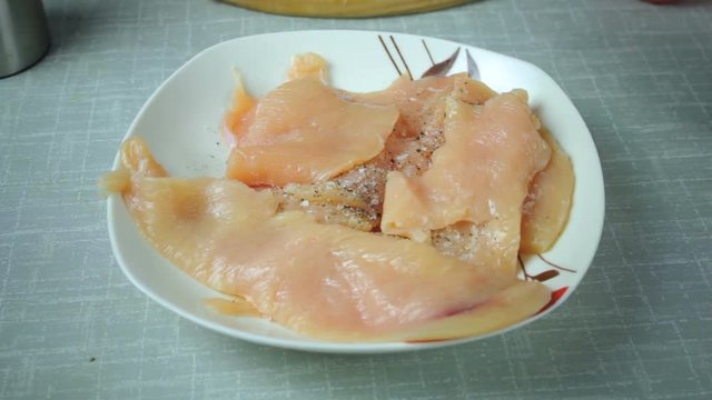 Cook adding spices, salt and pepper on the chicken. Raw chicken is cut into slices and lay on a plate.
