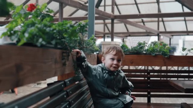 A cute little boy leaning on a fence with plants, sitting on a bench. Rain is falling. Slow mo