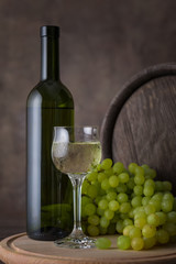 A bottle of white wine, a glass of white wine on a background of grapes and barrel on wooden table