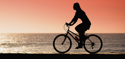Silhouette of young woman cyclist on sunset,along seashore summer beach