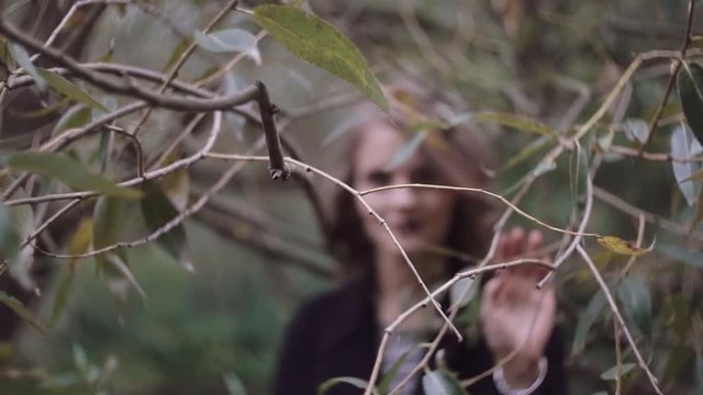 Stylish beautiful young blonde girl wades through the branches of trees, looking into the camera. slow motion