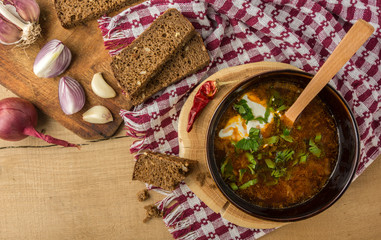 Tasty and nutritious borsch-soup with meat, potatoes, cabbage, tomatoes, beans, sour cream, parsley, onion, garlic, dark bread in a clay plate on a wooden tray and wooden table. Top view.