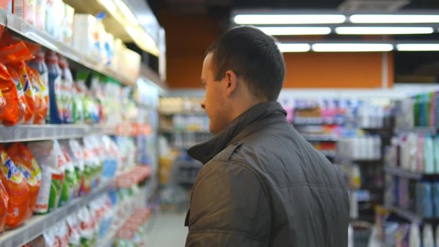Young man choosing household chemicals in supermarket. Handsome guy selects fabric softener or washing powder at the store and put it a shopping cart. Department of cleaning product and home care
