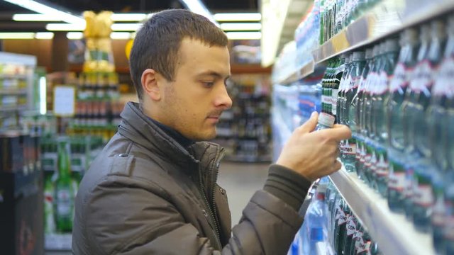 Young man choosing and buying bottle of mineral water at the supermarket. Guy taking product from shelves at grocery section of shop. Man buys drinks in store. Close up