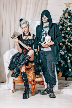 Young Gothic Couple Posing Near A Christmas Tree. Goth Style Male Model With Skull And Goth Girl With Axe In Christmas Decorations.