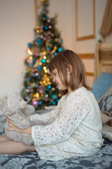child girl in  dress with toy bear on  bed near  Christmas tree