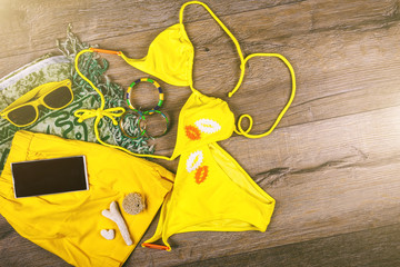 Set of beach clothes yellow bikini, bracelets, shorts, glasses on dark wooden background. Top view. Summer Holiday Concept.