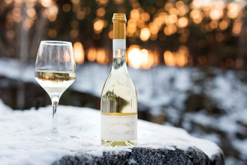 Love, romance, holiday, New Year celebration concept. Bottle and glass of white wine chilled by snow in winter forest on sunset.