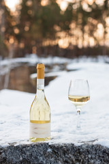 Love, romance, holiday, New Year celebration concept. Bottle and glass of white wine chilled by snow in winter forest on sunset.