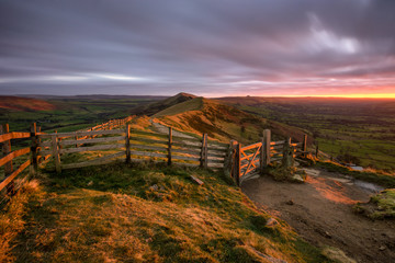 Glowing red light from the rising sun shining on countryside gate at Mam Tor in the Peak District with moody clouds.