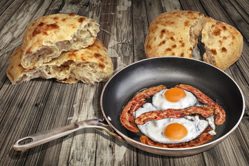 Sunny Side Up Eggs Fried with Bacon Rashers in Old Frying Pan Set on Weathered Garden Table With Two Torn Pitta Bread Loafs