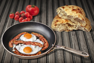 Sunny Side Up Eggs Fried with Four Bacon Rashers in Old Steel Frying Pan Set on Weathered Garden Table With Torn Pitta Bread and Bunch of Fresh Ripe Tomatoes