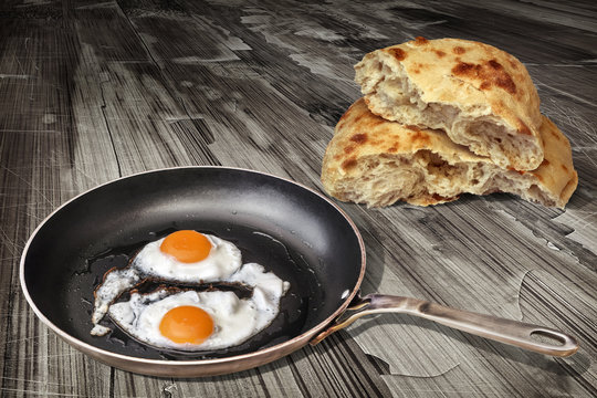 Pair of Fried Sunny Side Up Eggs in Old Heavy Duty Steel Frying Pan With Torn Pitta Bread Loaf Set on Weathered Cracked Flaky Garden Table