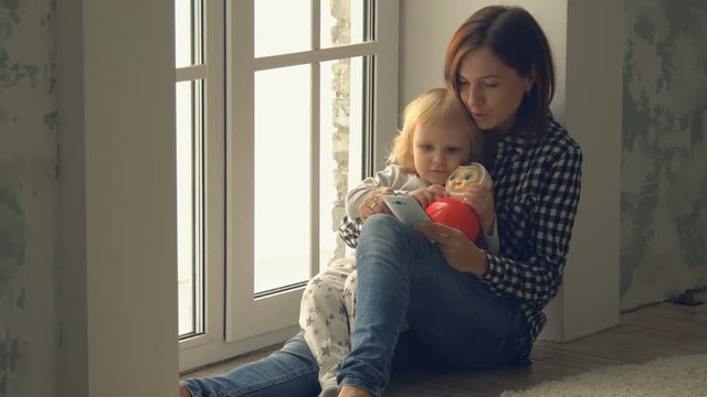 Mother with little daughter seats near the window and uses smartphone