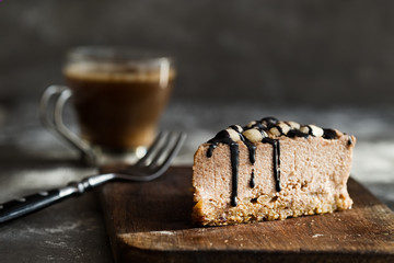Slice of raw chocolate mousse cake with cashew, hazelnuts and dark chocolate glaze topping and coffee on a wooden and grey background. Vegan sugar, gluten free dessert. Copy space