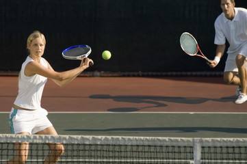 Young female tennis player hitting ball with doubles partner standing in background