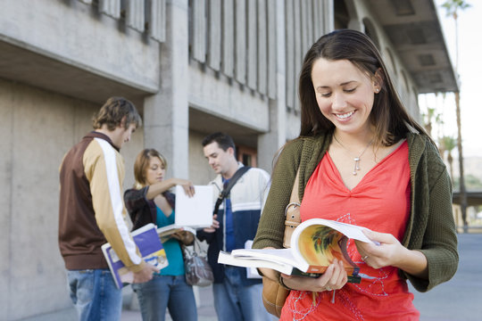 Young female student reading book with friends in background at campus