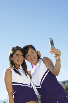Happy Asian cheerleader with teammate taking picture through cell phone