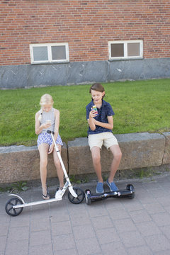 Sweden, Smaland, Anderstorp, Girl (12-13) with scooter and boy (14-15) with hoverboard sitting on wall and using phones
