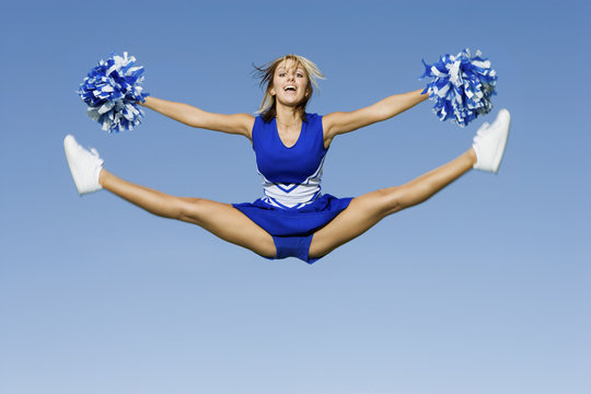 Full length of excited cheerleader with pompoms doing splits against blue sky