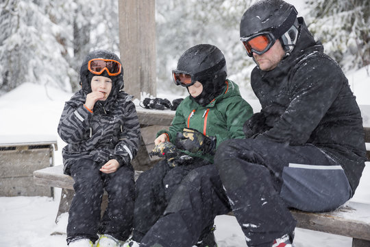 Sweden, Dalarna, Salen, Mature man and boys (6-7, 8-9) wearing helmets and goggles sitting on bench surrounded by winter landscape