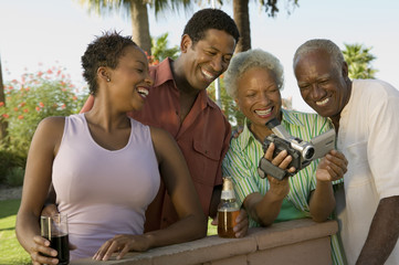 Senior couple and mid-adult couple looking at camcorder outdoors.