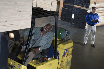 Happy senior male worker stacking wooden planks by forktruck with man writing on the clipboard