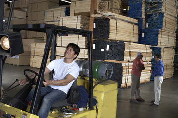 Fototapeta na wymiar Happy male worker driving forktruck in warehouse with people having conversation in the background