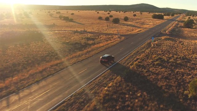 AERIAL: Black car driving along empty country road at golden summer sunset