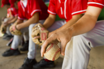 Low section of baseball team mates sitting in dugout with player holding a ball in foreground - obrazy, fototapety, plakaty