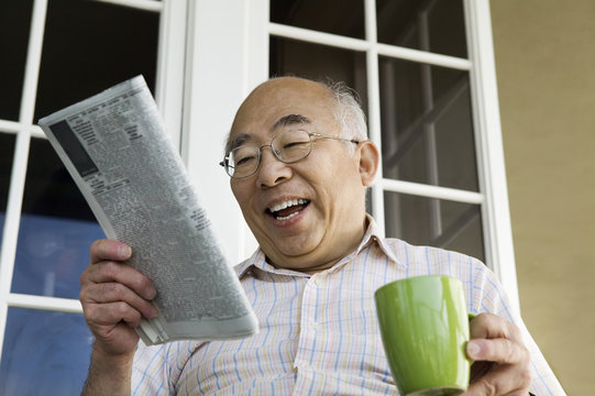 Cheerful senior man with coffee cup reading newspaper in front of house