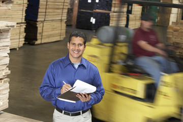 Portrait of a smiling confident man with clipboard in front of forklift in warehouse