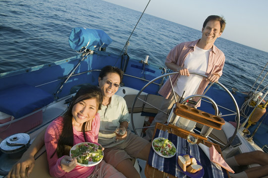 Happy Couple Having Food With Friend Standing By The Steering Wheel Of The Yacht