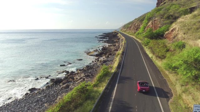 AERIAL: Red convertible car driving along coastal road above rocky shore