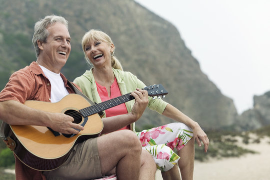 Middle Aged Man Playing The Guitar By A Cheerful Woman At The Beach