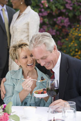 Cheerful mature woman feeding fruit to man with friends in background
