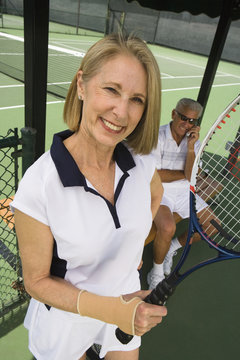 Portrait of a happy senior woman holding tennis racquet with man on call in background