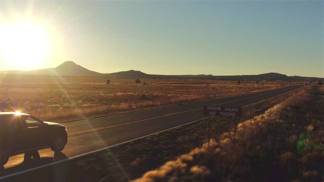 AERIAL: Black car driving along empty country road at golden summer sunset