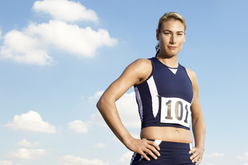 Portrait of a confident female athlete standing with hands on hip against the sky