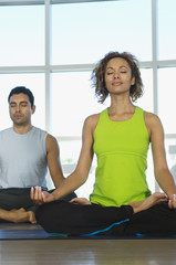 Middle aged woman sitting in lotus position with man in the background