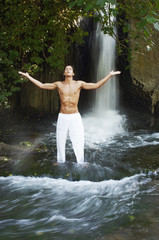 Young man with arms outstretched meditating while standing against waterfall