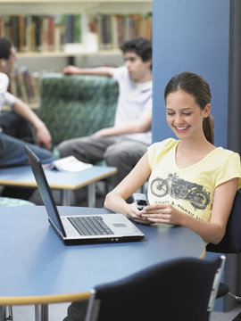 Happy teenage girl using cellphone with laptop on table in library