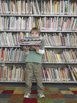 Full length of happy young boy holding stack of books in library