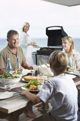 Family sitting around table eating barbecued food with mother standing at grill grilling
